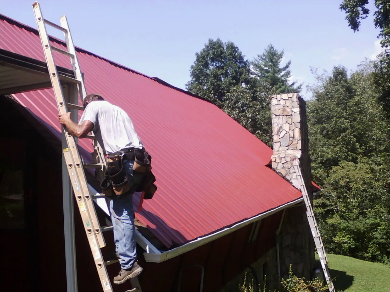 Roofer on a ladder beside a red metal roof and stone chimney — Soffit & Fascia Repair by Roofing Brothers in Chadwicks